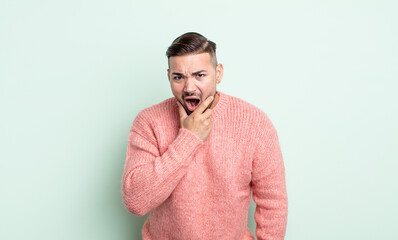 young handsome man with mouth and eyes wide open and hand on chin, feeling unpleasantly shocked,...