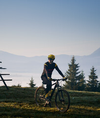 Man in cycling suit riding bicycle on grassy hill in the morning. Male bicyclist enjoying the view of majestic mountains during bicycle ride. Concept of sport, bicycling and nature.