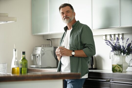 Happy mature man drinking coffee at home in the kitchen, enjoying hot drink in the morning on weekend.