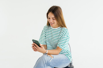 Naklejka premium Portrait of woman sitting on white chair and texting on the phone typing something with excitement on white background