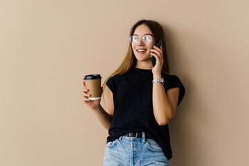 Beautiful excited young pretty woman posing isolated over beige background wall holding mobile phone talking drinking coffee.