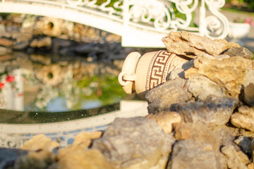 The decorative bridge and jug are reflected in the artificial pond. Beautiful summer photo of decorative garden decoration.
