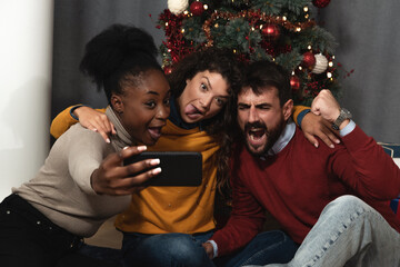Three young friends celebrate Christmas and New Year's Eve together by joking and taking selfies with a smartphone goofy and making funny facial expressions in front of the Christmas tree