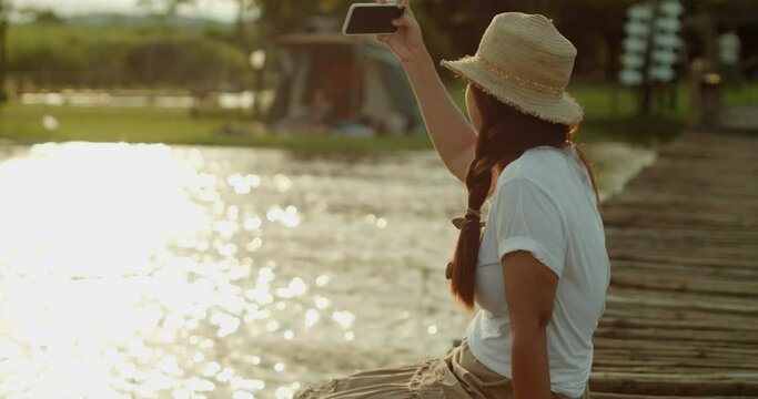 A female tourist in a brown hat, white T-shirt and brown skirt sits on a wooden bridge by the lake, use her mobile phone take the photos relaxing and joyfully in the evening at the sunset time.	