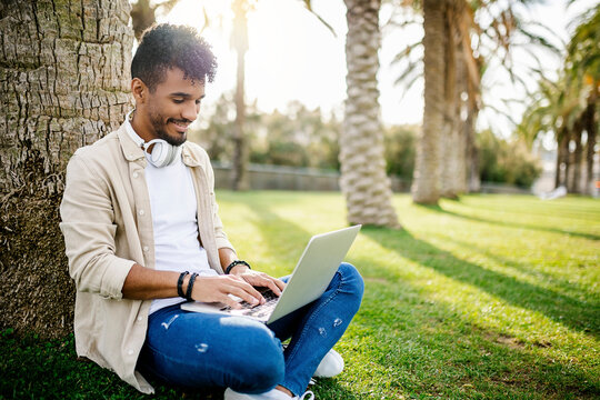 Smiling young man using laptop while sitting by tree trunk