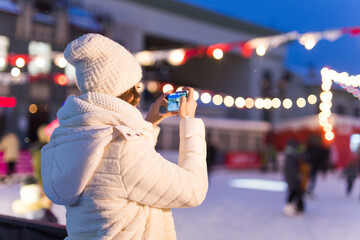 A woman on the rink is skating and taking selfie on smartphone. New Years Eve and Christmas. Fairy lights. Ice and snow mood concept. Winter sport.
