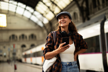 Young woman waiting for the train standing at the railway station. Beautiful girl using the phone while waiting for the train.