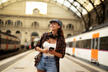 Young woman waiting for the train standing at the railway station. Beautiful girl using the phone while waiting for the train.