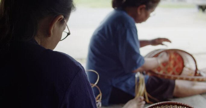 Rack focus shot of 2 Thai female farmers weave basket in traditional Thai art patterns to sell as extra income in countryside of Thailand.
