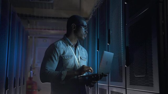 Engineer network administrator at work upon checkup. African-American man spbas with glasses types on notebook computer keyboard next to servers cabinets closeup