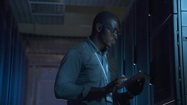 Engineer serves present data center. Serious African-American guy spbas with glasses does testing of server equipment using tablet bright screen in center closeup