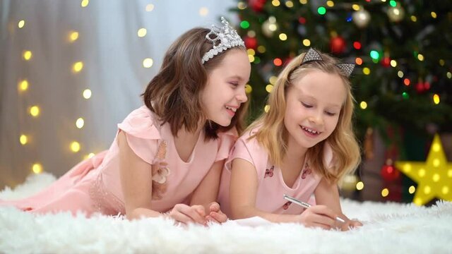 beautiful and happy little girls in pink dresses write a letter to santa claus at the Christmas tree. the relations of the sisters in the family. Happy New Year. fashionable elegant clothes for girls.