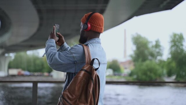 Young man taking selfie photo with smartphone and standing on city bridge spbas. American African guy holds phone in his hand and looks with happy smile, takes pic or has online talk and stands by