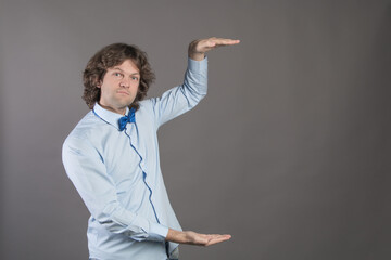 Horizontal shot of handsome Caucasian man with bristle, shows height of something with both hands, pretends holding item or thing, demonstrates size, wearing shirt and bow-tie, isolated on grey
