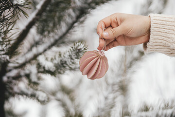Hand in cozy sweater holding modern bauble on background of pine tree branches in snow. Decorating...