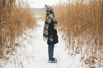Stylish hipster woman standing among dried grass at snowy winter lake. Atmospheric calm moment. Tranquility. Young female in cozy stylish clothes relaxing in snowy park. Wanderlust
