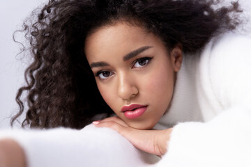 Confident multiracial young woman wearing white sweater, lying with folded hands, studio portrait. Contemporary curly mixed race female with perfect makeup posing on photoshoot.