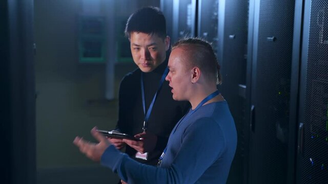 Male programmers discussing business and using tablet while checking networks on render farm spbas. Close-up view of two young men talk with smiles and examine equipment, use device in hands and stand