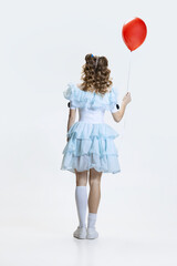 Back view of young girl wearing Halloween dress, costume of movie character standing with red balloon in hand isolated over blue background