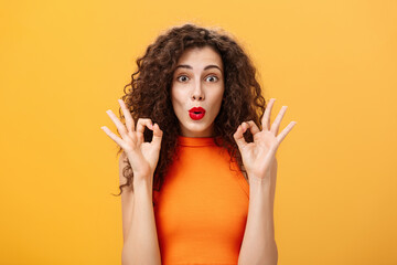 Enthusiastic charming caucasian girl with curly hairstyle in red lipstick and orange top showing okay gesture being intrigued and delighted folding lips approving and agreeing on awesome adventure - obrazy, fototapety, plakaty