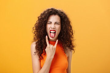 Carefree rebellious stylish urban female with curly hairstyle sticking out tongue yelling from satisfaction and joy showing rock n roll sign chilling and enjoying cool concert over orange background