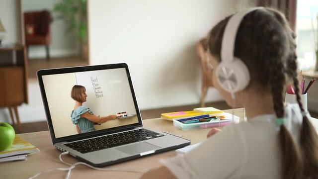 Online education at home. Little girl pupil with modern headphones listens to teacher at English video lesson spbas spbd via notebook at table backside view
