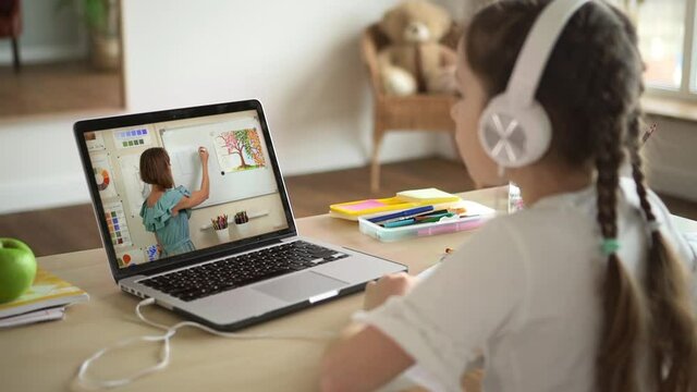 Online education at home. Little girl with long plaits learns to paint with skilled teacher on screen at art lesson by video call at table spbas spbd backside view