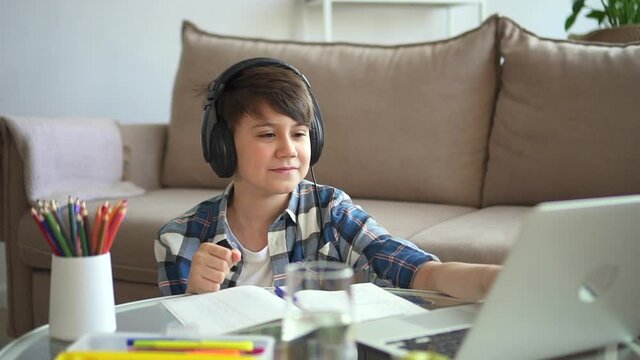 Boy school student greet teacher, wave hand for tutor and start online lesson Spbd. Smiling elementary student sit by sofa with laptop virtual leaning. Concept homeschooling, notebook, headphone