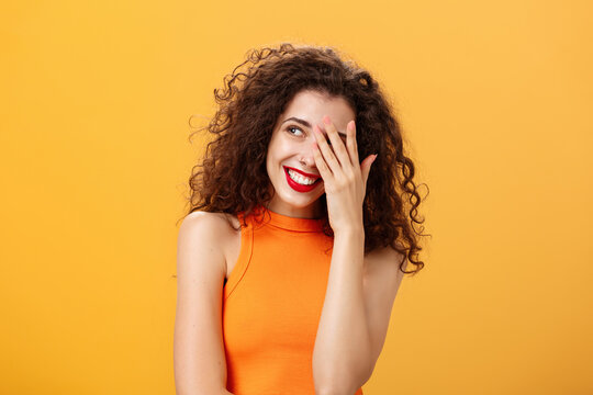 Woman being praised by coworkers feeling awkward and shy hearing congratulations with received award covering face peeking left with joyful satisfied smile posing over orange background
