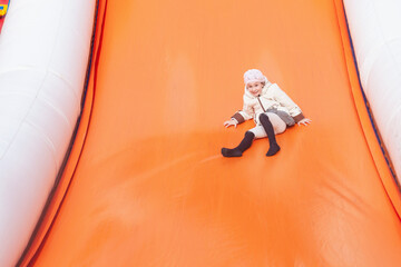 a girl in a light hat and jacket slides down the orange slide of a rubber trampoline