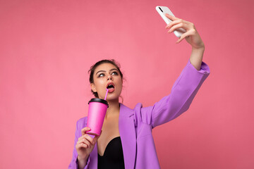 Close-up portrait of a beautiful young brunet woman in fashionable purple suit on a pink background in studio in a holding a popular phone and taking selfie photo and drinking through a straw beverage