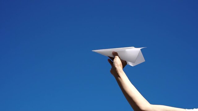 白い紙ひこうきを飛ばす女性の手元 青空背景