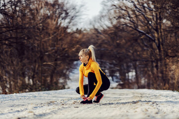 Sportswoman crouching on snowy path in nature at winter and tying her shoelace. Sportswear, winter...