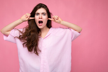 Naklejka premium Portrait of young emotional winsome pretty curly brunet woman with sincere emotions wearing casual pink shirt isolated over pink background with copy space and covering ears with fingers and yelling