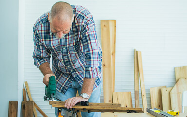 Senior old Caucasian man wearing check shirt, apron, making DIY wooden furniture, using equipment to drill woods with happiness while standing alone at indoor home. Retirement and Hobby Concept