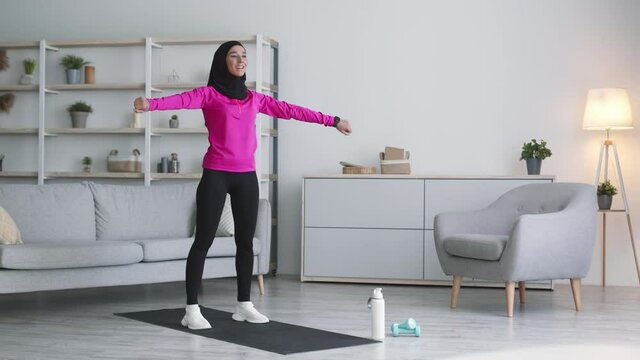 Domestic gym. Young positive muslim lady wearing sportswear and hijab exercising at home, warming up her hands