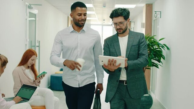 It S A Very Busy Day At The Office, Two Working Men In Suits Are Walking Throughout The Halls And Talking While Holding A Tablet, They Pass By A Lot Of Different People