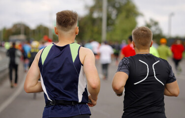 Young men in sportswear running marathon in city, wireless earphones in their ears, other contestants on blurred background. Runners at sports competition