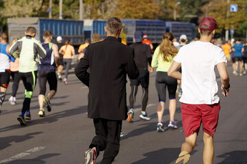 Obraz premium Businessman in black office suit running city race with other people and recording voice message on smartphone. Marathon contestants. Concept of striving