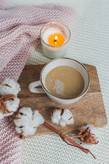 Autumn flat lay. A pink knitted blanket, a cup of hot cappuccino, candle light, a wooden stand, a white pumpkin, a branch of cotton.