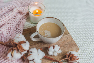 Autumn flat lay. A pink knitted blanket, a cup of hot cappuccino, candle light, a wooden stand, a white pumpkin, a branch of cotton.