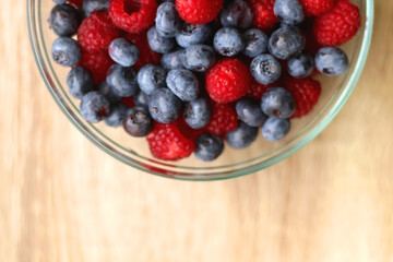 Glass bowl filled with raspberries and blueberries on wooden table. Top view.