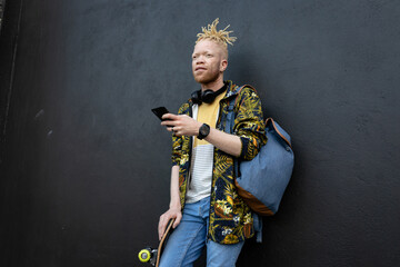 Thoughtful albino african american man with dreadlocks holding skateboard using smartphone
