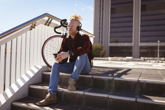 Thoughtful albino african american man with dreadlocks wearing headphones using smartphone