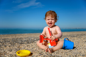 Little laughing girl sitting on the beach and playing with colorful toys. Summer beach vacation concept.