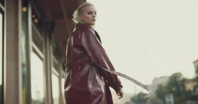 Back view of blond woman walking through an empty city center. Lady turns around, looks at camera then starts to run. Stylish person in red leather coat, morning urban scene.