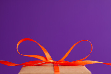 Close up of brown gift box with a orange satin ribbon bow on the bottom on vibrant purple blurred background with copy space. Holiday autumn and Halloween concept.