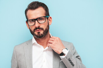 Portrait of handsome confident stylish hipster lambersexual model.Sexy modern man dressed in elegant suit and spectacles. Fashion male posing in studio near blue wall. Looking at camera in glasses