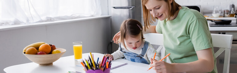 Parent and child with down syndrome drawing on paper near orange juice and fruits in kitchen, banner.