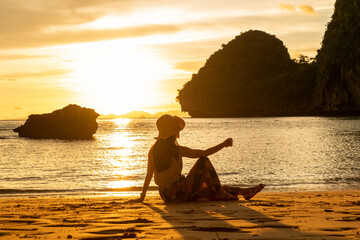 female tourist in a hat sits on the beach at sunset, relaxing during the holidays. 